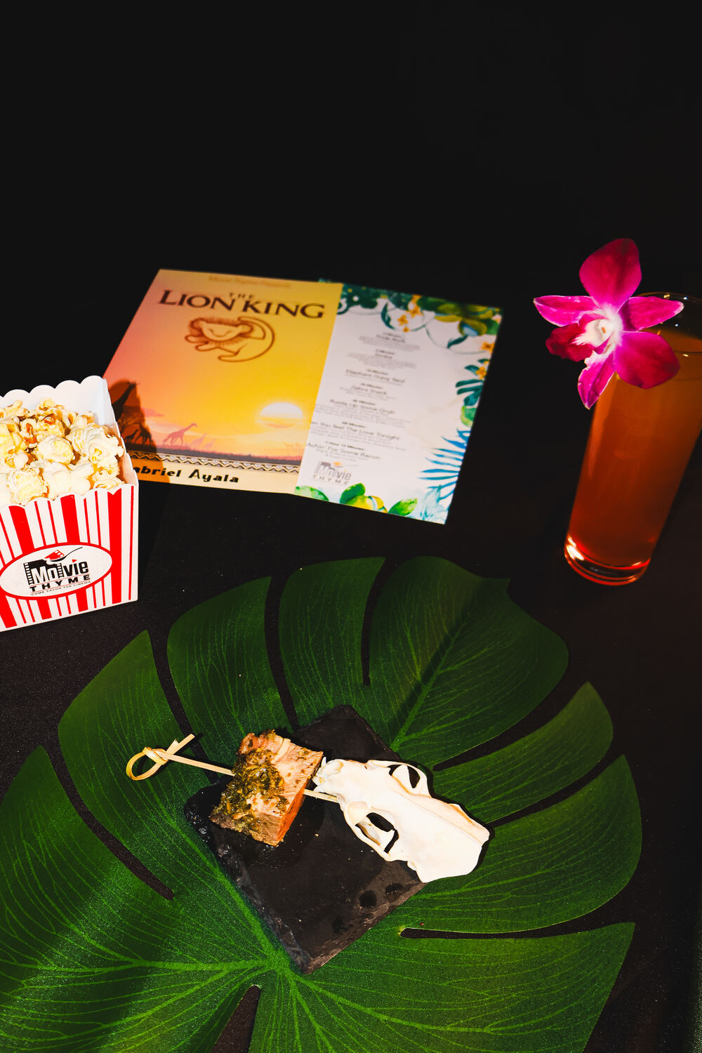 A Lion King course at Movie Thyme: a glazed salmon skewer on slate over a monstera leaf, beside a branded striped popcorn box, a printed Lion King menu card and a tall tropical cocktail with an orchid