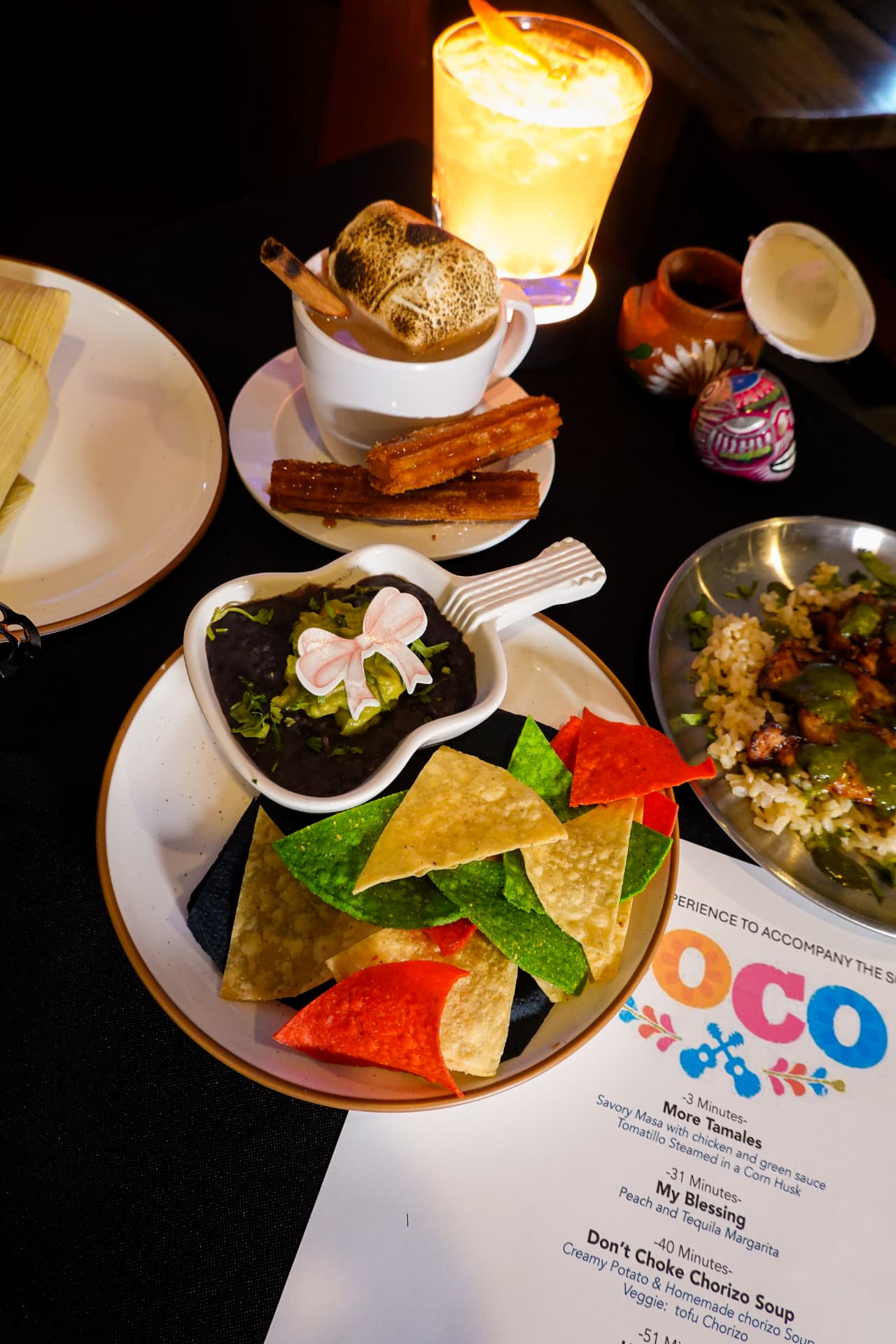 A Coco night at Movie Thyme: corn-husk tamales, tortilla chips in Mexican flag colours with guacamole and black beans in a guitar-shaped dish, churros beside a marshmallow-topped hot chocolate, and a printed timed Coco menu card